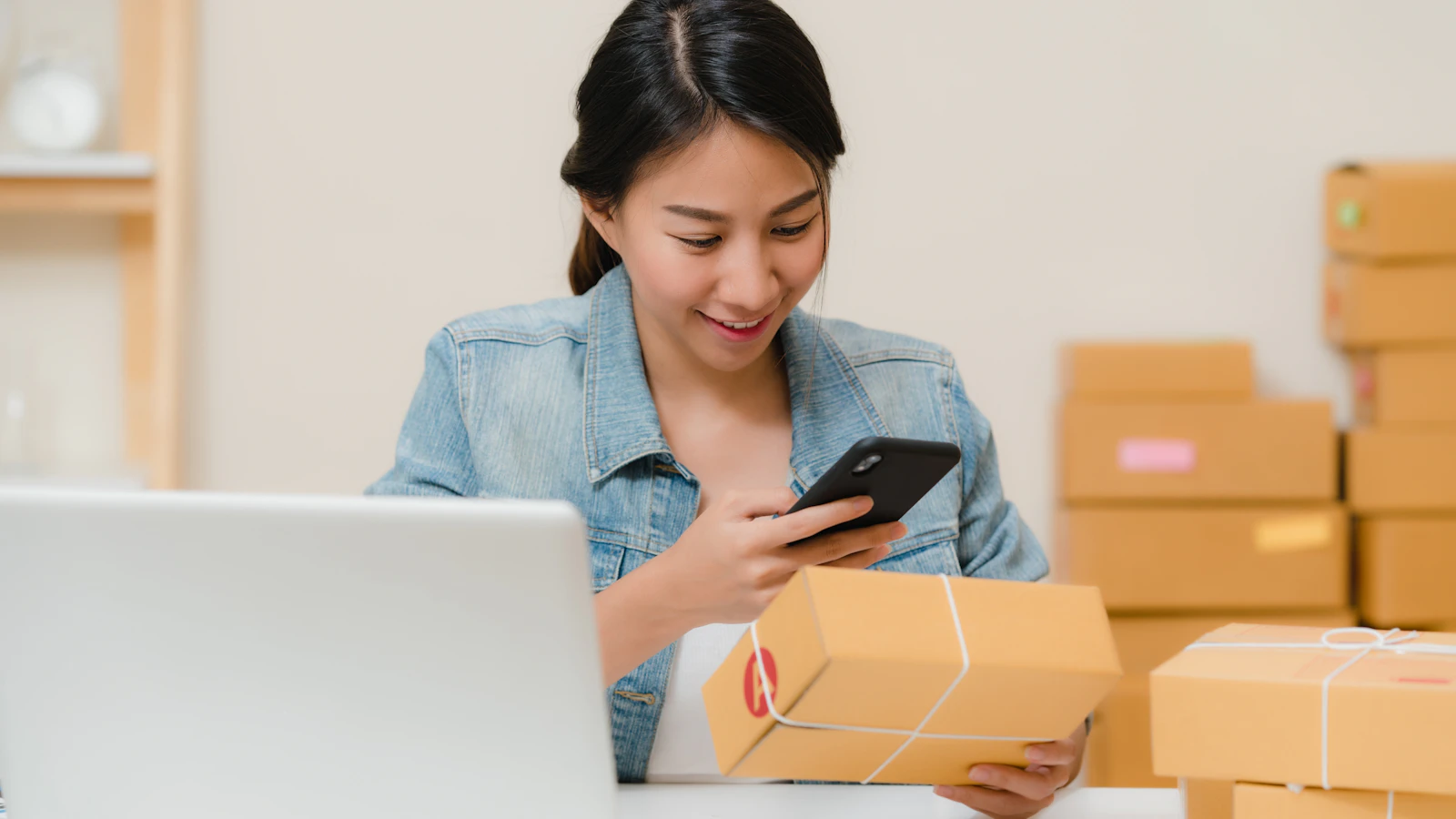 Commerçante vérifiant un colis avec son téléphone près de cartons de stock